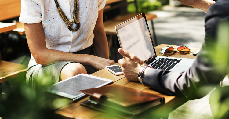 Uma mesa com vários materiais em cima, um laptop e duas pessoas conversando